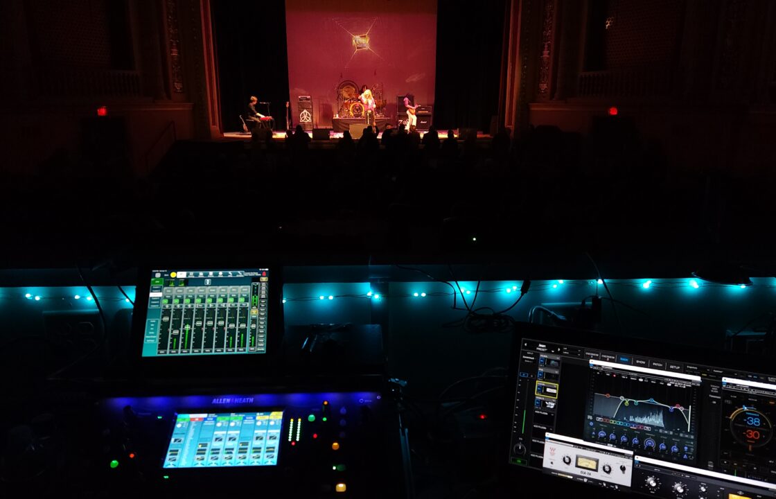 Mixing board and laptop set up in the sound booth in front of a band playing on an orange lit stage