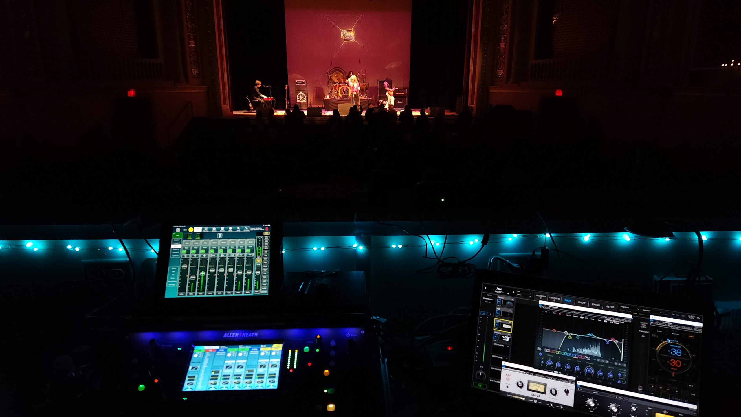 Mixing board and laptop set up in the sound booth in front of a band playing on an orange lit stage