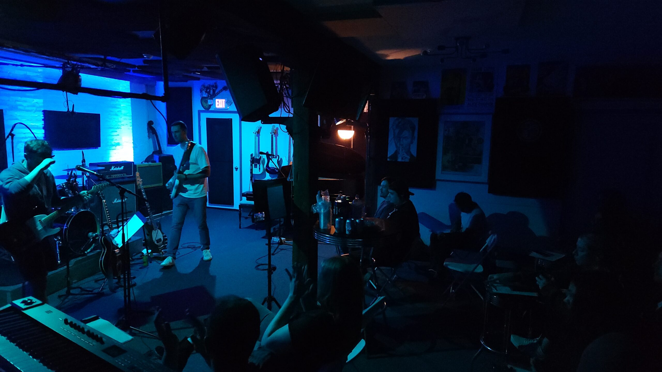 A band performing in our studio with only our blue back lighting on. Two additional people can be seen listening at our high top table.